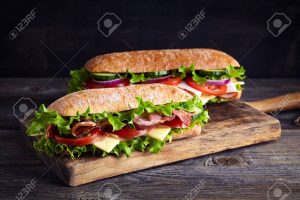 Two fresh submarine sandwiches with ham, cheese, bacon, tomatoes, lettuce, cucumbers and onions on wooden cutting board
